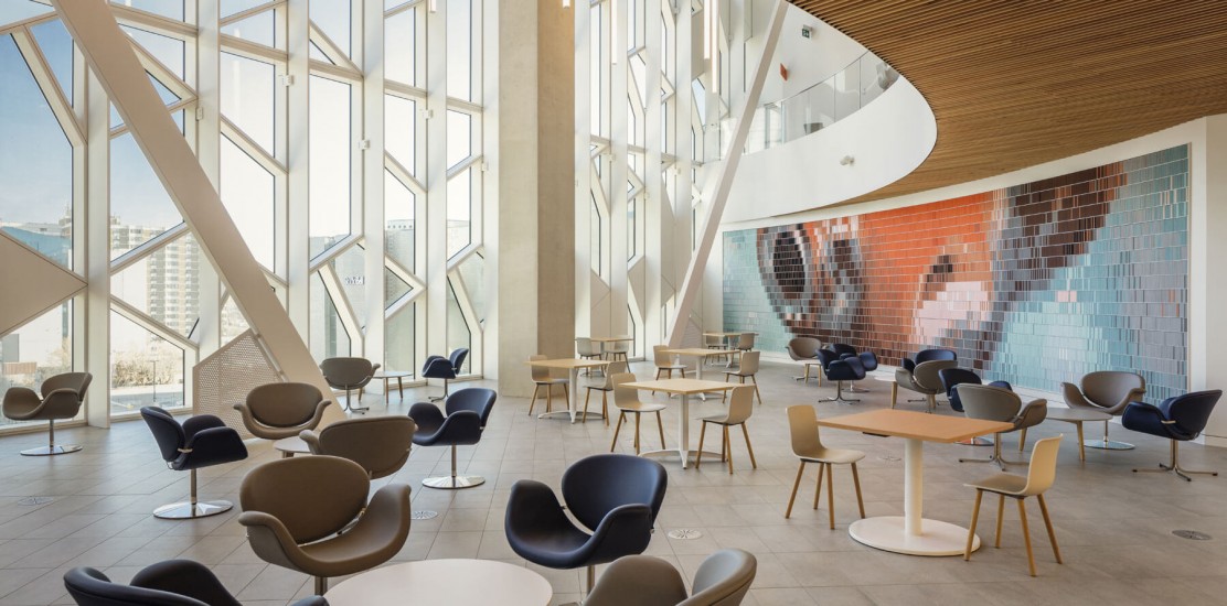 Central Library interior with floor-to-ceiling windows, a curved wooden ceiling, and a large colorful mural on one wall. The seating area includes comfortable armchairs, small tables, and a few high-top tables.