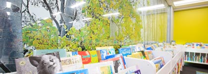 Children's picture books in shelves, behind them is a mural of a tree. 