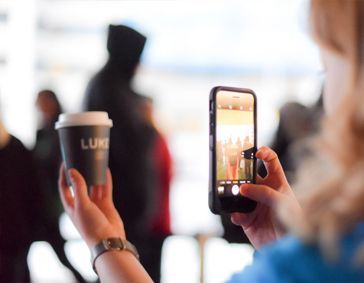 background image: Photograph of a person taking a photo of a to-go coffee cup with their smart phone. 