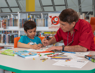 A child and an adult sitting at a table working on a colouring sheet together. 
