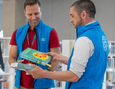 Two Library staff members wearing blue vests review books together at a service desk inside the library.