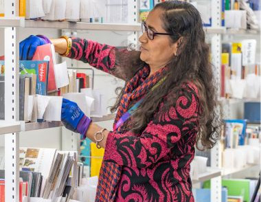 A Library staff member shelves books in a library collection area, wearing blue gloves and reading glasses.