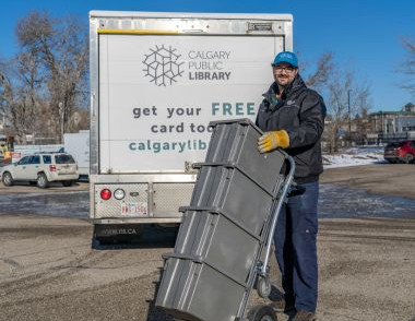 A Library staff member loads grey book bins onto a dolly beside a Calgary Public Library delivery truck in a parking lot on a winter day.