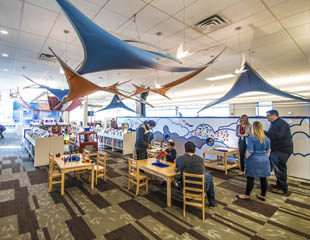 An Early Learning Centre that has sail like fabric across the top of the ceiling. Below it is tables with chairs and people sitting and doing crafts.
