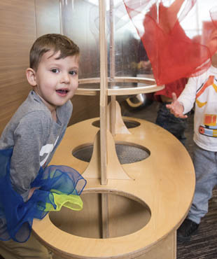 A little boy leaning forward on a structure. They are throwing silk fabric in the air that is being blown around by the machine.