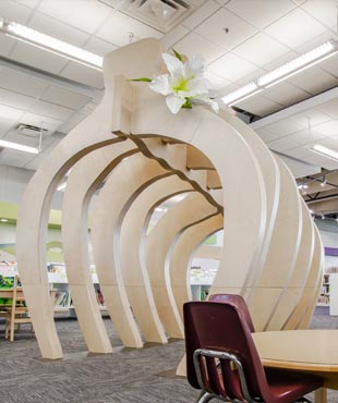 A wooden structure resembling the ribcage of a dinosaur is large enough for people to walk under. On top of the structure, there is a white and green flower.