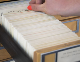A person's hand with a red fingernail browsing through index cards in a wooden library card catalogue drawer.