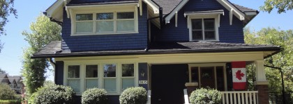 A blue house with white trim, a gabled roof, and a Canadian flag hanging on the porch.
