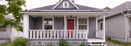 A small gray bungalow with white trim, a covered porch with a red front door, and a lattice skirt around the base.