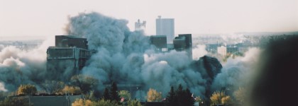 A cloud of dust and debris engulfs a building as it is demolished, with the city skyline visible in the background.