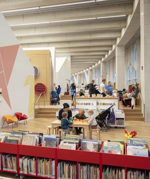 This is a picture of the Early Learning Centre in Central Library. The left side of the photograph has pink origami swans on the wall. A family is sitting at the table playing with blocks, and behind them, you can see other patrons browsing the books.