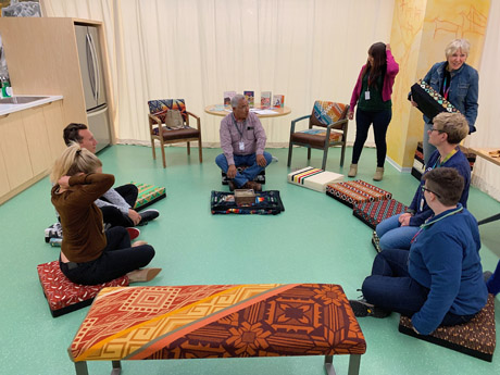 This is a visual of seven people sitting on cushions in a circle on the floor of the Indigenous Language Resource Centre. They are chatting with an Indigenous Elder.