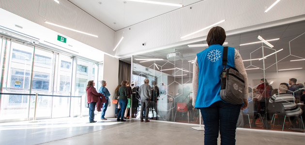 A person wearing a blue vest with the Calgary Library logo on the back walking into a meeting room. There is a line of people on the left side walking into the same meeting room.