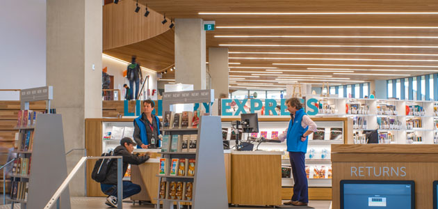 This is a picture of the inside of Central Library, showcasing the 'new' 'new' 'new' bookshelves. In the background, you can see the front desk with a staff member in a blue vest helping a patron.