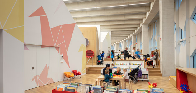 This is a picture of the Early Learning Centre in Central Library. The left side of the photograph has pink origami swans on the wall. A family is sitting at the table playing with blocks, and behind them, you can see other patrons browsing the books.
