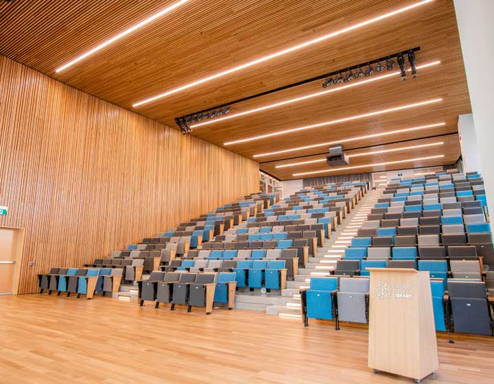 background image: An image of Patricia A. Whelan Performance Hall. The image has a podium on the right-hand side with rows of blue, gray and black chairs in the background in an auditorium style. 