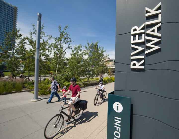background image: A photograph of the Jack and Jean Leslie RiverWalk. In the picture, you can see people riding their bikes and a dad holding his son's hand and walking.