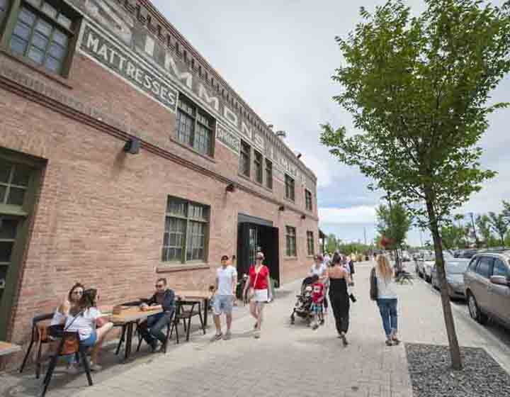 background image: This picture is of the Simmons Building, which is close to the RiverWalk. In front of the building, people are walking, and three people are sitting down chatting and enjoying lunch. 