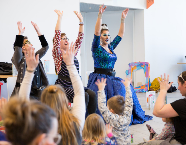background image: A storytelling session in a public space features three drag performers dressed in an elegant blue gown, a purple striped dress and, a trench coat with a gold mask on and a tiara reading a children's book to a group of children and adults.
