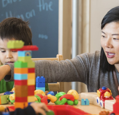 A photograph of an adult with a group of children. She is helping them stack blocks. 