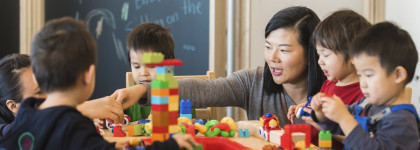 A photograph of an adult with a group of children. She is helping them stack blocks. 