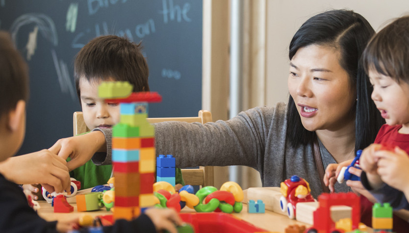 A photograph of an adult with a group of children. She is helping them stack blocks. 