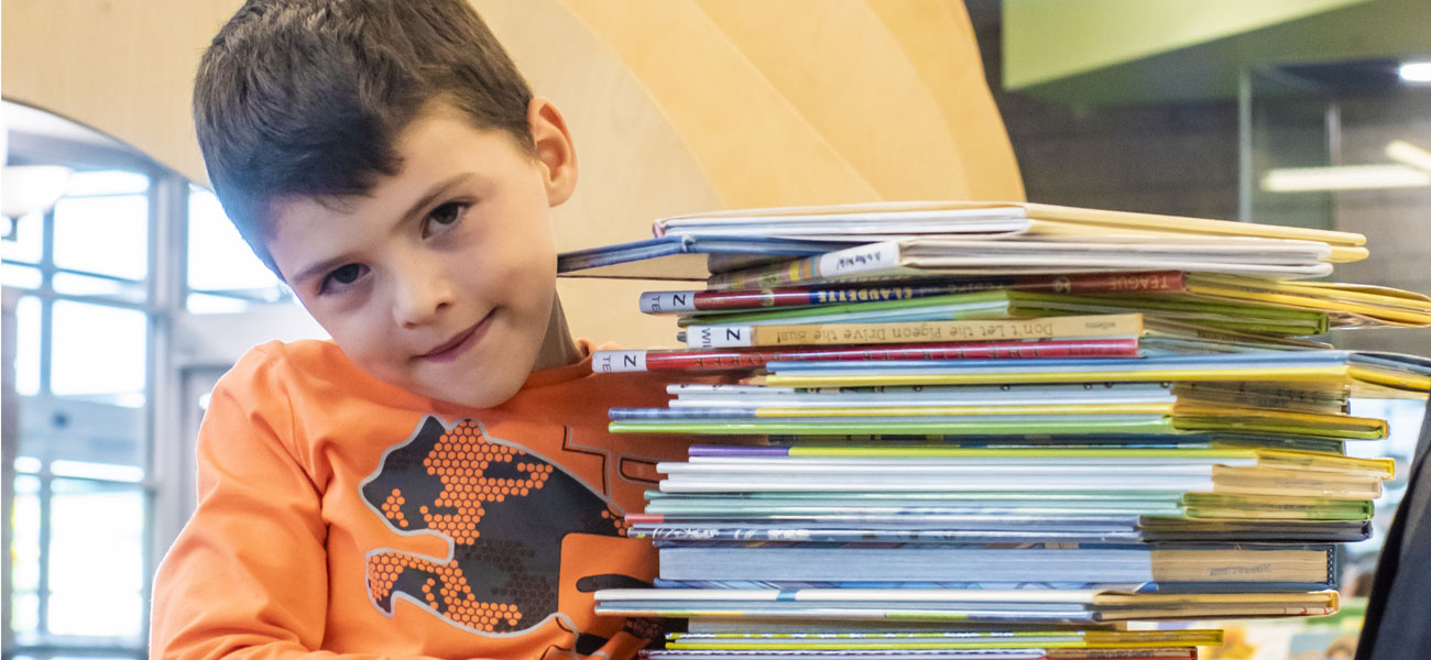 A kid holding a stack of books and is looking at the camera. child
