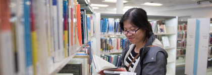 A person standing in front of bookshelf, reading a novel. 