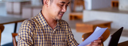 A adult male sitting at a desk, holding a piece of paper in front of him.