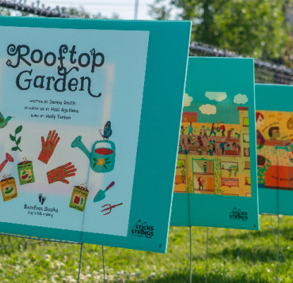 A row of large, illustrated book pages from the children’s book Rooftop Garden is displayed outdoors along a fence, creating a “storywalk” experience. The pages, mounted on teal boards, show colorful drawings of gardening scenes and tools. The grassy area