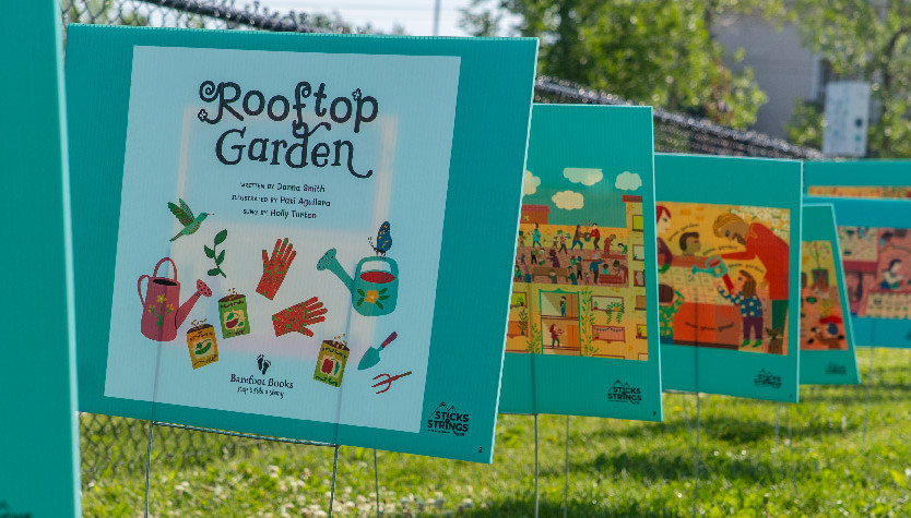 A row of large, illustrated book pages from the children’s book Rooftop Garden is displayed outdoors along a fence, creating a “storywalk” experience. The pages, mounted on teal boards, show colorful drawings of gardening scenes and tools. The grassy area