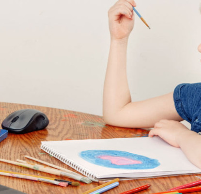 A child sitting at a wooden table with paint supplies and a laptop around them, as they follow along with an online art tutorial.