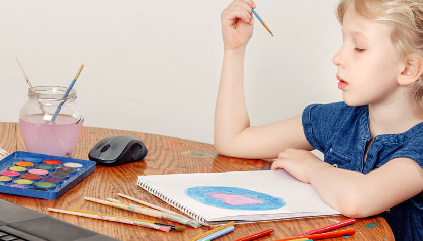 A child sitting at a wooden table with paint supplies and a laptop around them, as they follow along with an online art tutorial.