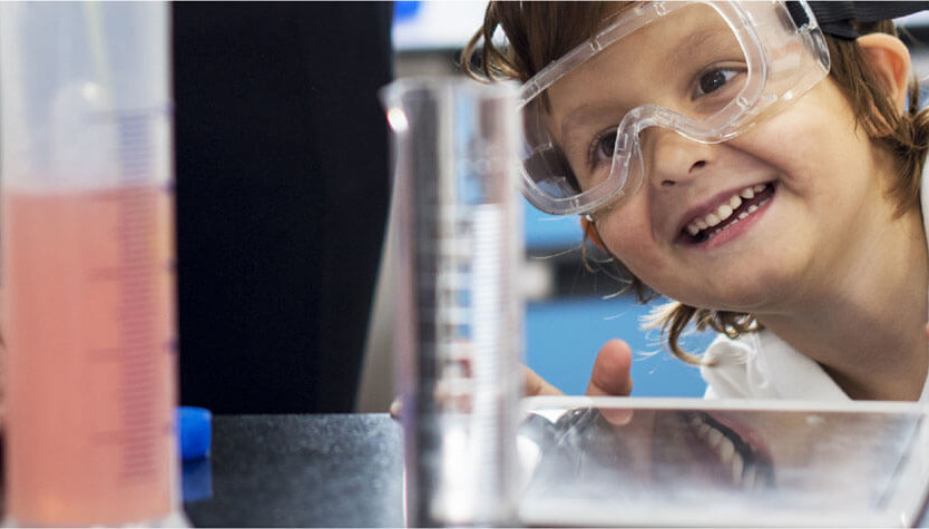 A child dressed up as a scientist in a lab coat looking at science lab equipment.