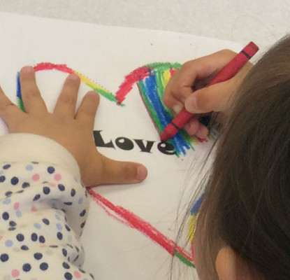 A little girl in a polka-dot jacket is drawing a heart on a piece of paper. In the centre of it is the word love. She is colouring the heart with rainbow colours.