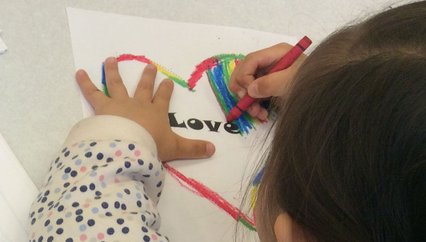 A little girl in a polka-dot jacket is drawing a heart on a piece of paper. In the centre of it is the word love. She is colouring the heart with rainbow colours.