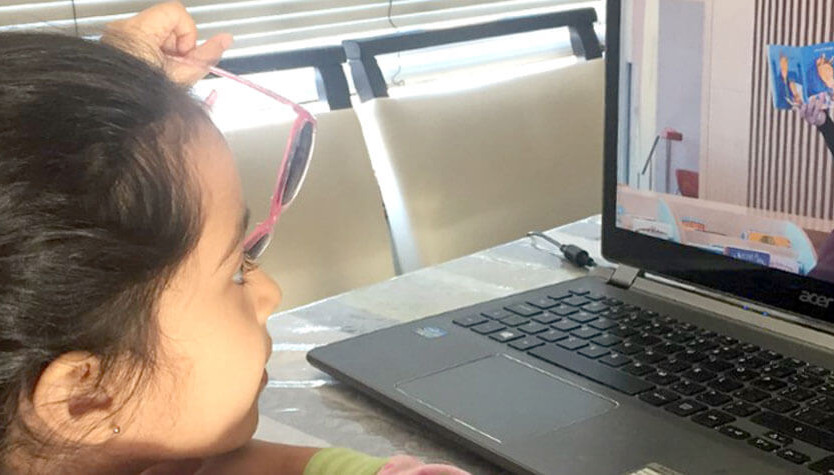 Photograph of a child sitting in front of a laptop screen. They are watching an online storytime program