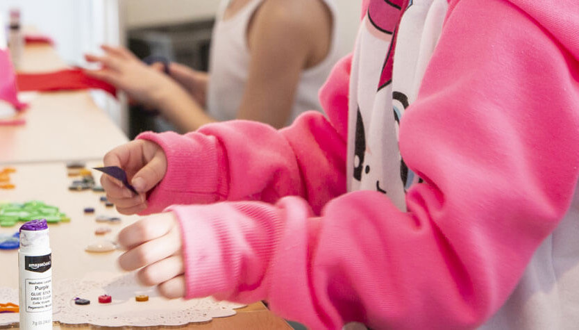 Photograph of a child's arms and hands. They are wearing a white and pink sweatshirt and they are doing a craft. 