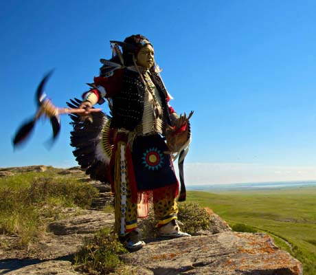 A Indigenous dancer, adorned in vibrant regalia with feathers and intricate beadwork, stands on a rocky outcrop overlooking a vast prairie landscape. They hold a feathered staff aloft.