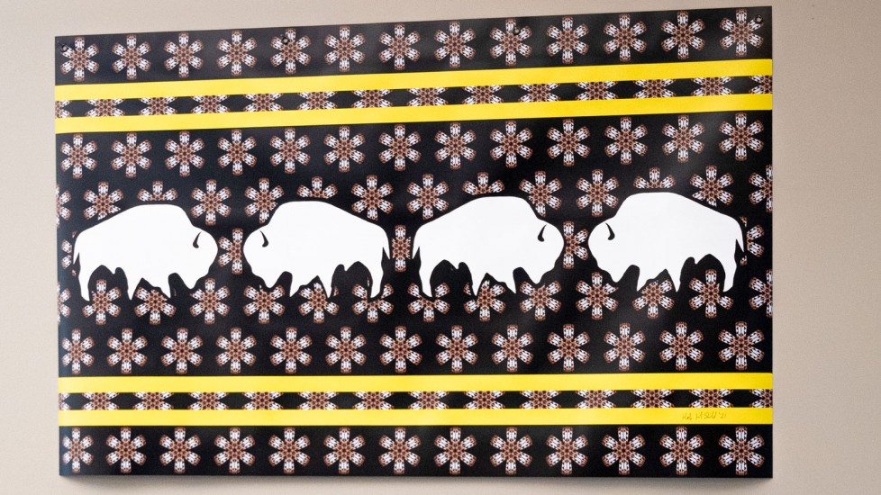 A piece of artwork featuring three white bison on a black background. The background is filled with a repeating pattern of stylized flowers and yellow stripes, possibly referencing Indigenous beadwork designs.