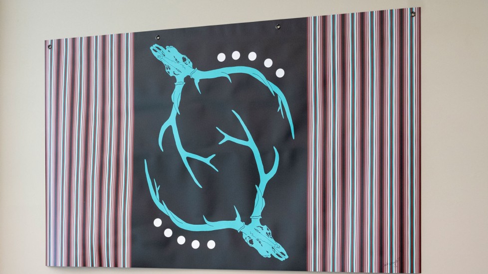A banner with a black center featuring two turquoise deer skulls with antlers, positioned mirror-image to each other. The background is filled with vertical stripes in turquoise, and maroon, evoking a ribbon shirt design.