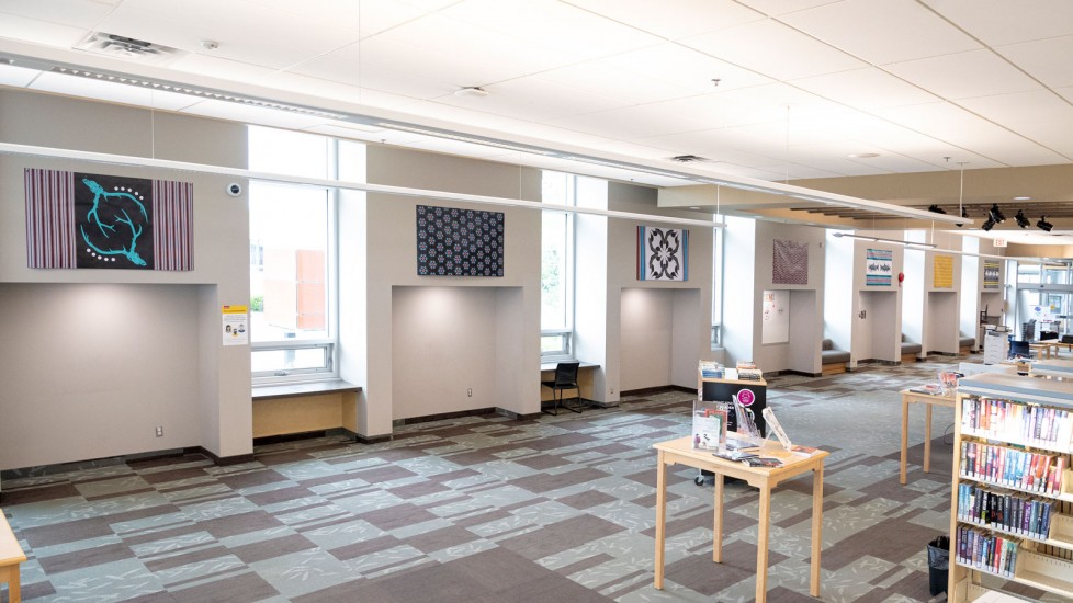A bright Library space with large windows and row of artwork displayed on the wall. Tables with reading materials and bookshelves are visible in the background.