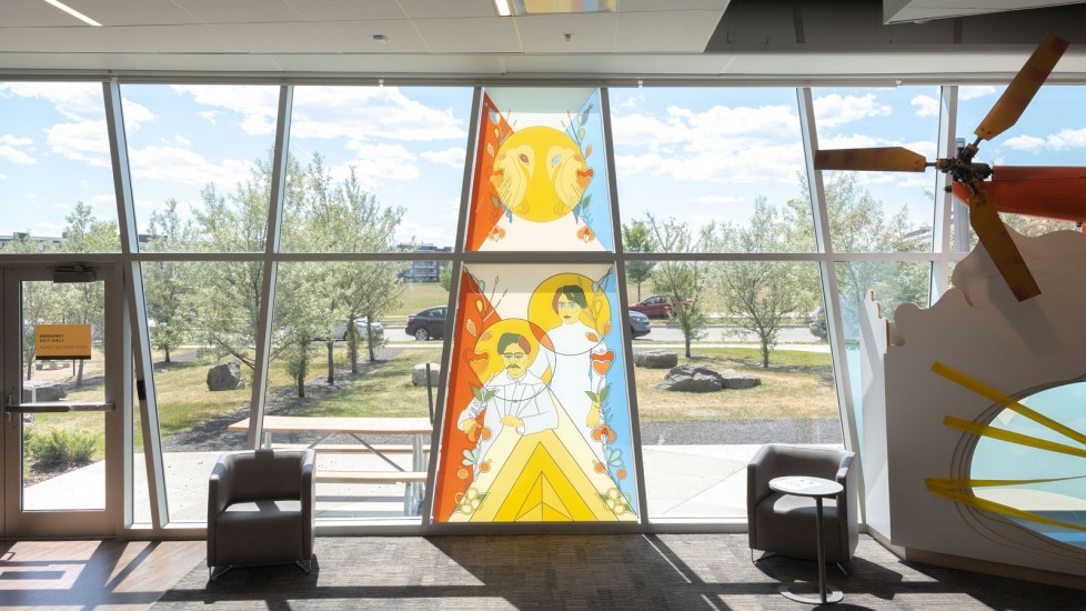  A modern Library interior with large windows showcasing a colourful art piece. The artwork features stylized figures and geometric shapes in a stained-glass style.