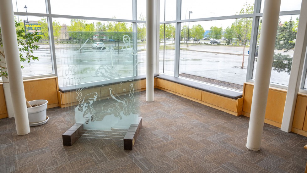 Glass panel etched with an image of otters swimming. The panel stands in a corner with windows, benches and potted plant nearby.