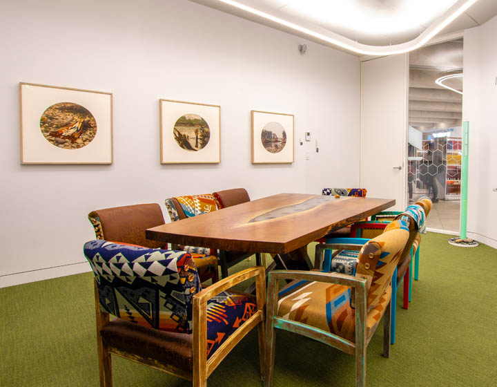 background image: This is a visual of the Elders' Guidance Circle. In the photograph, there is a long table in the centre, surrounded by chairs with embroidered patterns. Behind it, there are three different photographs hanging on the wall. 