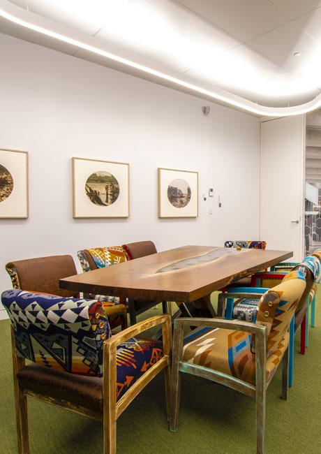A modern boardroom features a long wooden table with a unique, textured inlay and chairs upholstered with colourful Indigenous-inspired patterns. Three framed photographs hang on the white wall.