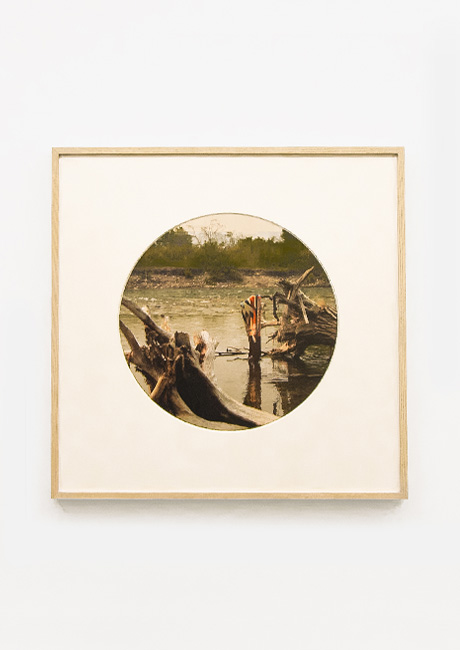 An oval-shaped photograph in a light wood frame shows a tranquil river scene. Driftwood and weathered wooden posts emerge from the calm water, hinting at a forgotten structure.