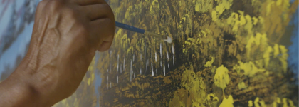A photo of a hand holding a paintbrush and painting a forest landscape.