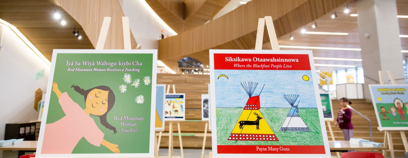 Two children's books, propped on easels, stands in the foreground of a bright, modern Library space. The book covers feature colourful illustrations. one depicting a young girl and the other a teepee and deer.