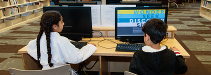 A girl and a boy are seated in front of computers in a library.